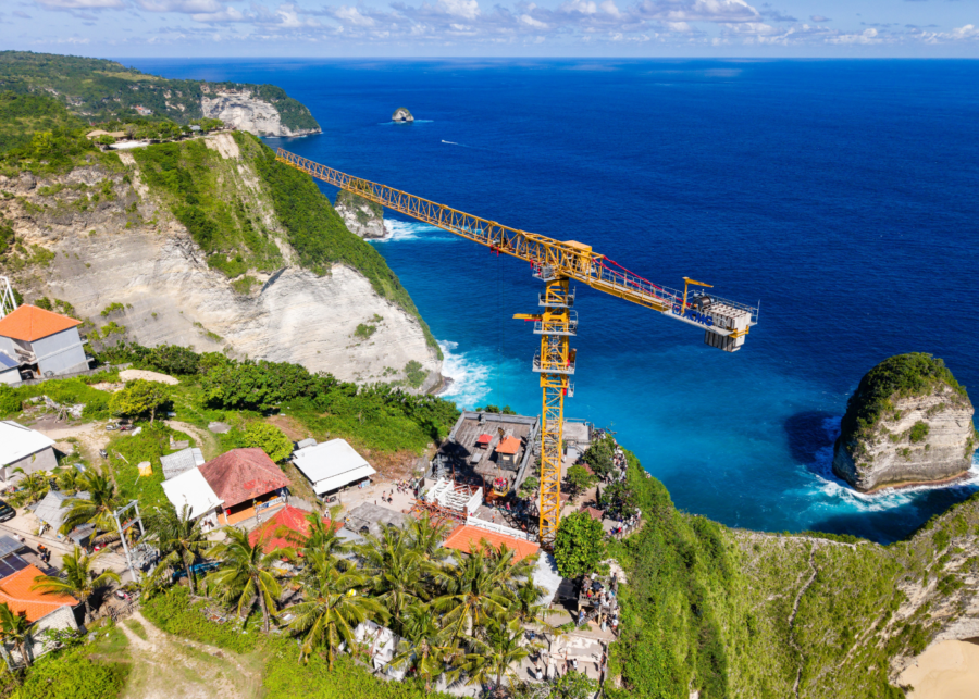 Aerial view of construction site for the glass elevator at Kelingking Beach, Nusa Penida, with a yellow crane and iconic cliff formation above turquoise waters.