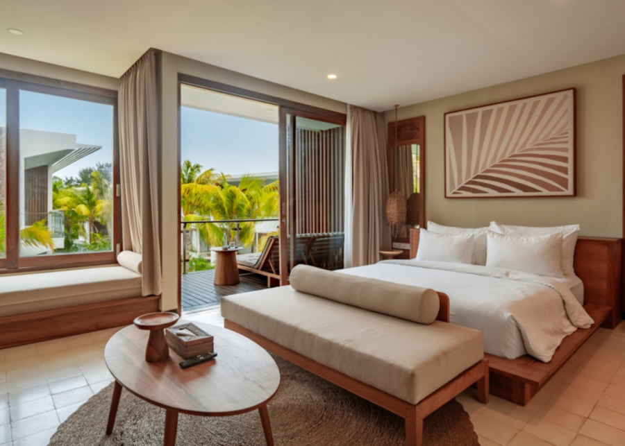 A bright, modern bedroom at Cocana Resort Gili Trawangan, featuring wooden furniture, a large bed, lounge seating and sliding doors leading to a balcony surrounded by palm trees.