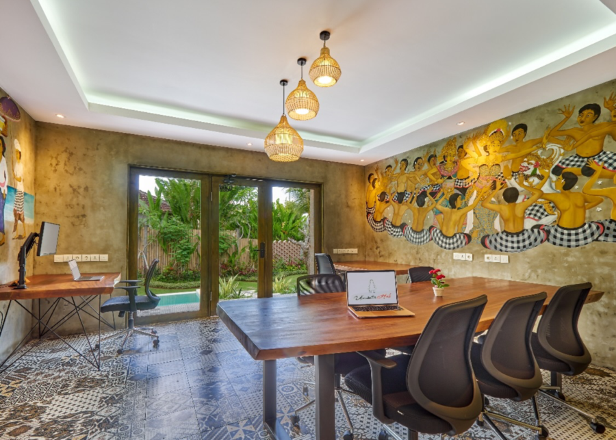 Meeting room at Uluwatu Hub coworking in Pecatu, with a long wooden table, ergonomic chairs, a traditional Balinese mural and pool views through glass doors.