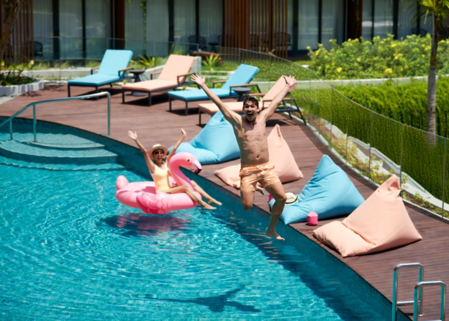 A man jumping into the pool and a child floating on a pink flamingo pool float at Aloft Bali Kuta at Beachwalk, with blue and peach bean bags and sun loungers on the poolside deck.