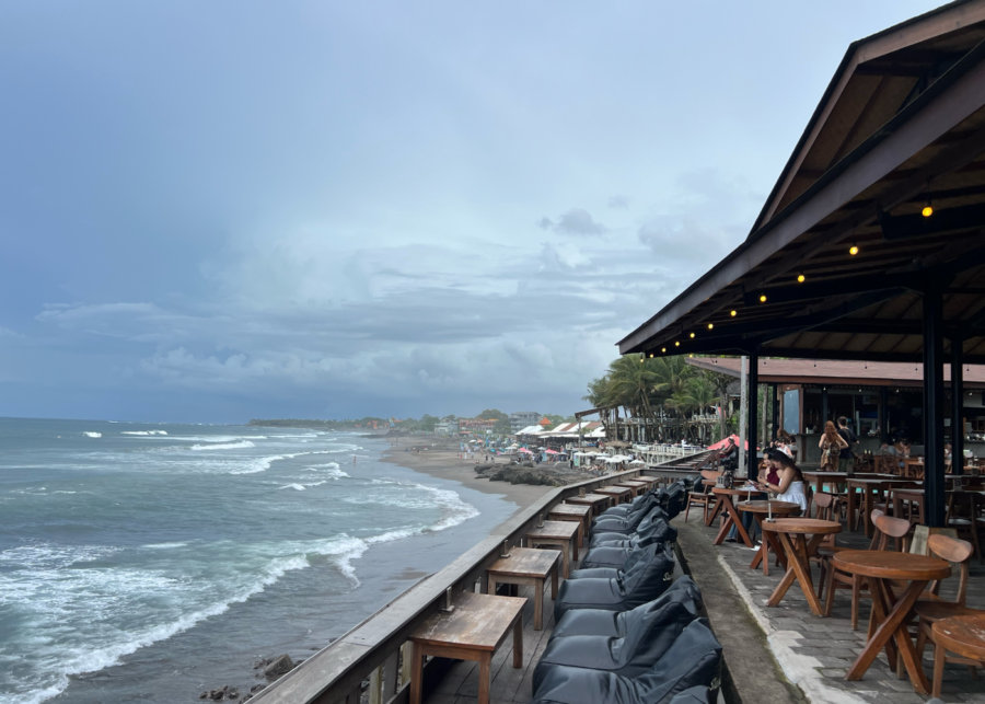 Oceanfront dining at Echo Beach Canggu with wooden tables, beanbags along the edge, and waves rolling in under a cloudy sky.