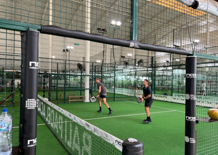 Indoor padel court at Bali Padel Academy Canggu with players mid-game, glass walls and green turf.