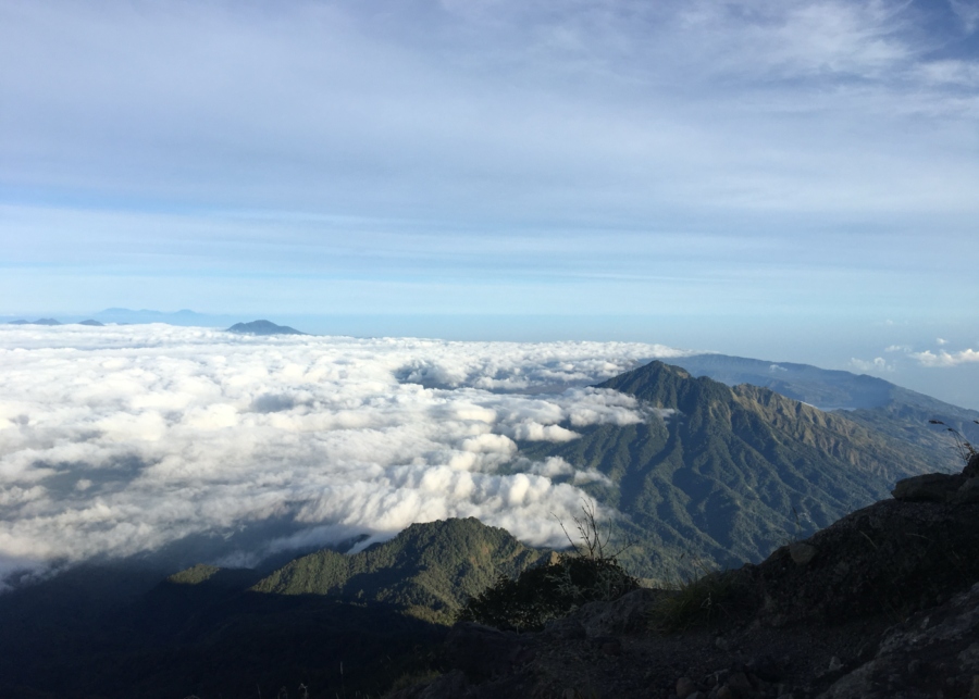 Sunrise view from Mount Agung summit above a sea of clouds, with rugged volcanic ridges and soft morning light breaking through the sky