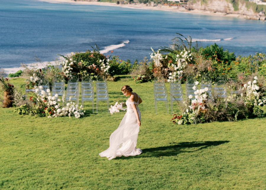 Bride walking through a clifftop garden wedding setup at Tirtha Bali, surrounded by white floral arrangements and clear chairs overlooking the ocean.