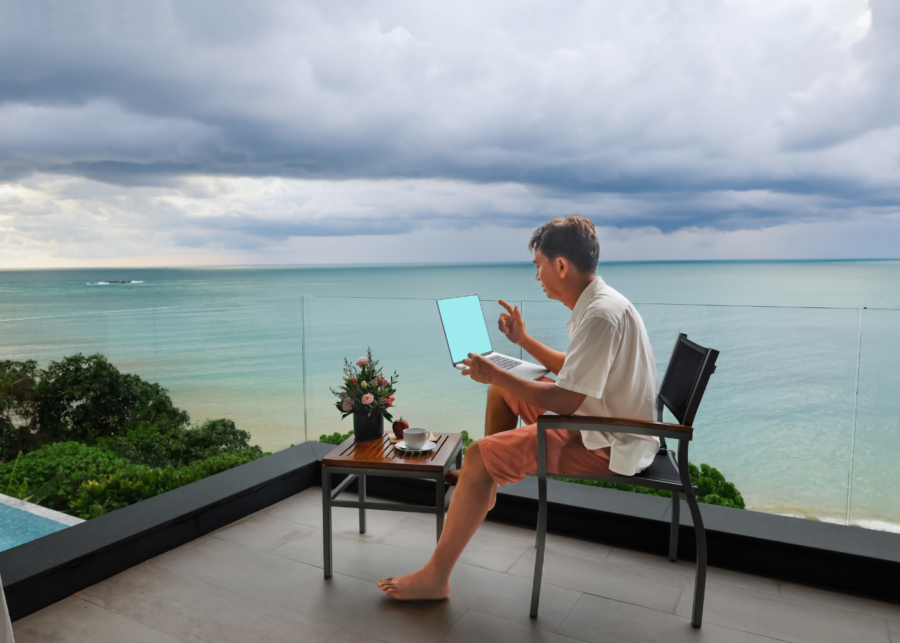 A man working on a laptop at an outdoor table on a clifftop terrace in Bali, overlooking the sea.