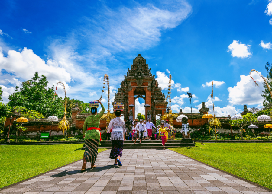 Two people in traditional Balinese dress walking towards a temple gate under a bright blue sky in Bali.