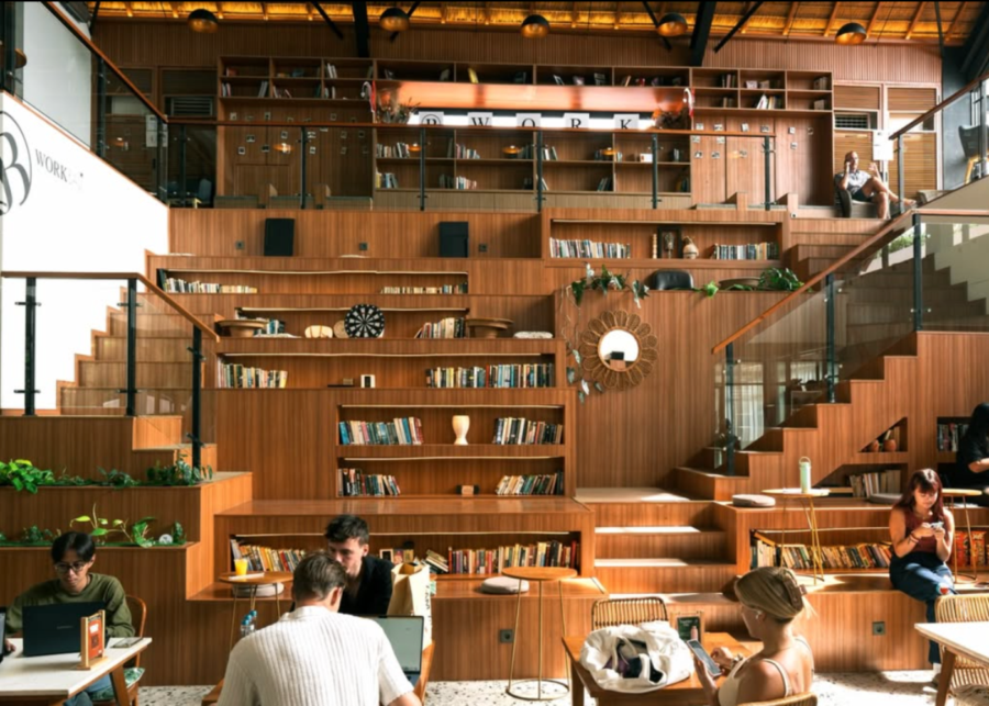 The multi-level wooden staircase library at BWork in Canggu, lined with books and plants, with people working on laptops at tables below.