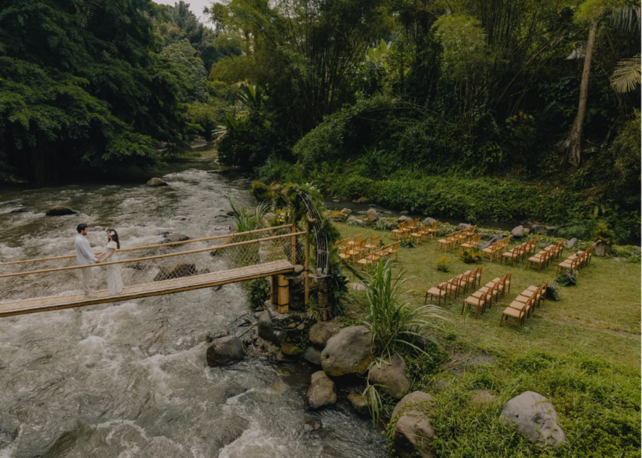 Bride and groom crossing a bamboo suspension bridge over the Ayung River at Bambu Indah, with wedding ceremony chairs set up on the riverside lawn, Ubud, Bali