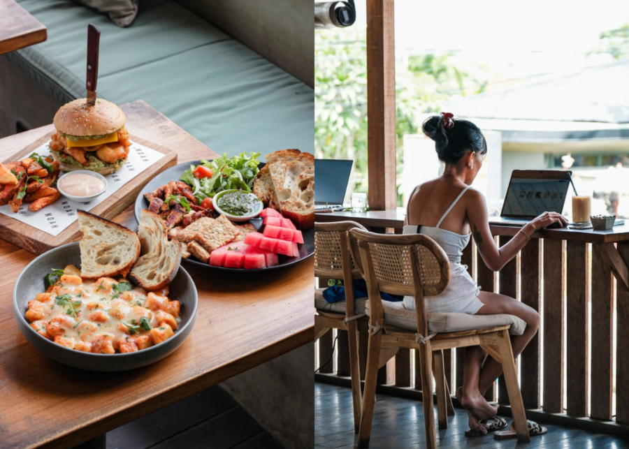 A woman working on a laptop at a wooden balcony bar overlooking greenery at NOAH Cafe in Pererenan, beside a spread of burgers, gnocchi, salad and fresh bread.