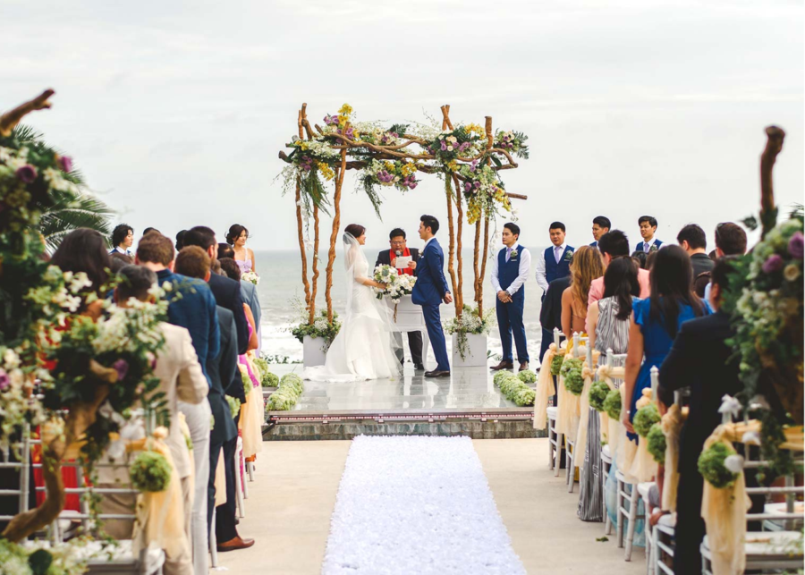 Bride and groom exchanging vows under a wooden floral arch at an oceanfront wedding ceremony, guests seated along a petal-strewn aisle, Soori Bali, Tabanan