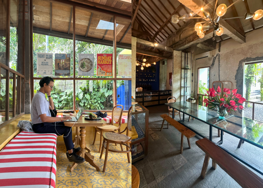A man working on a laptop on a striped bench by big windows onto greenery at Powder Room by R4D in Ubud, beside a rustic room with a glass-topped table, benches and a vase of red flowers.