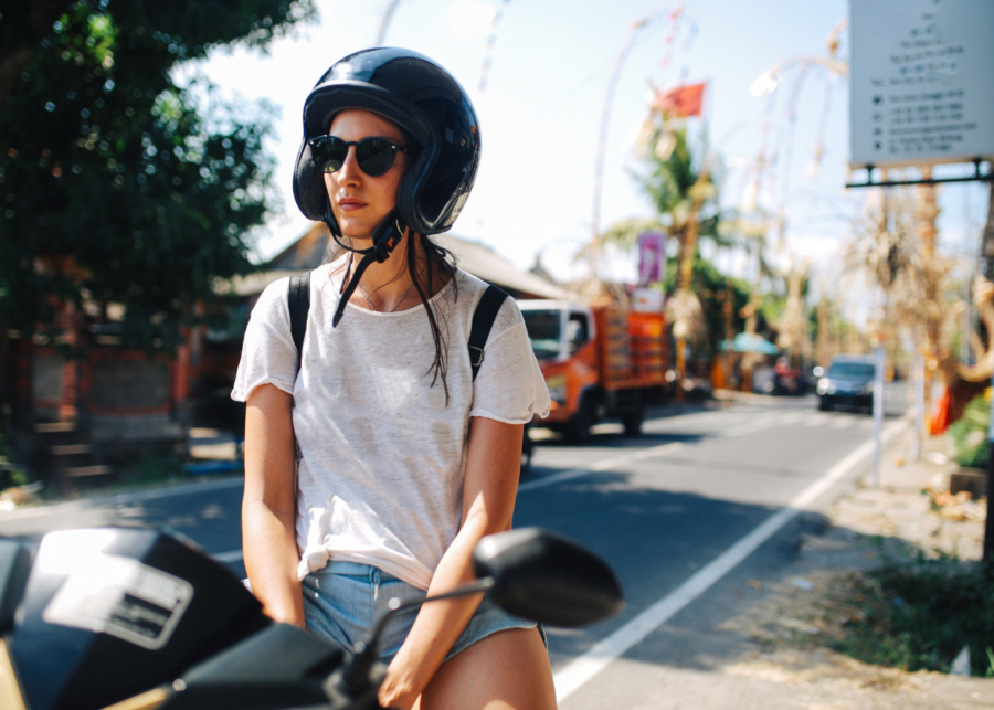 A woman wearing a helmet and sunglasses standing beside a scooter on a Bali street lined with palm trees and traditional penjor.