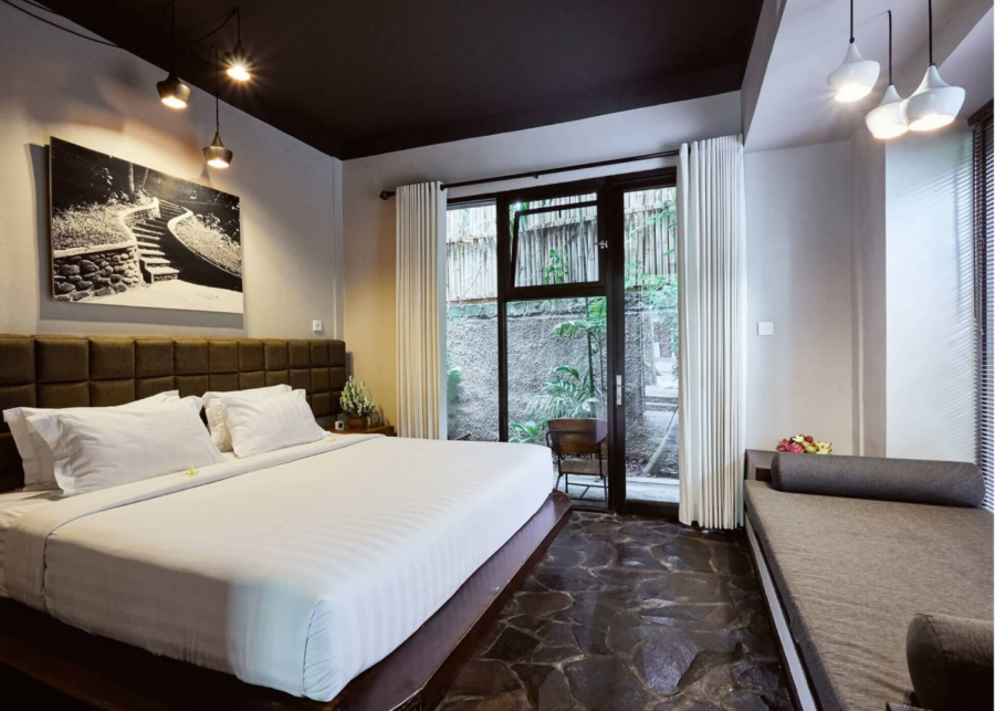 A private room at Outpost coliving in Ubud, Bali, with a dark ceiling, stone floors, a daybed and a glass door opening onto a garden.
