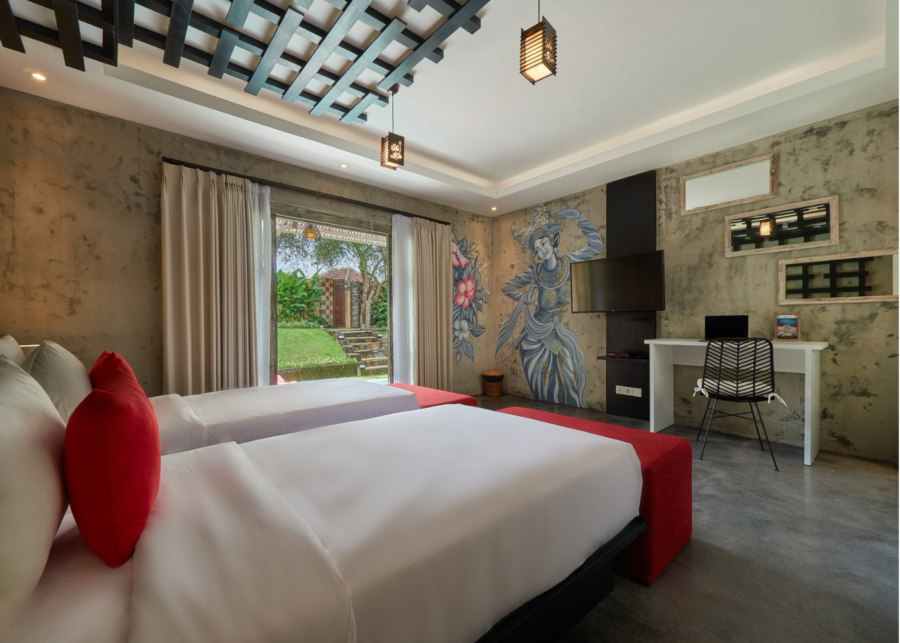 A twin room at Uluwatu Hub coliving in Uluwatu, Bali, with a Balinese dancer mural, a work desk and a door opening onto the garden.