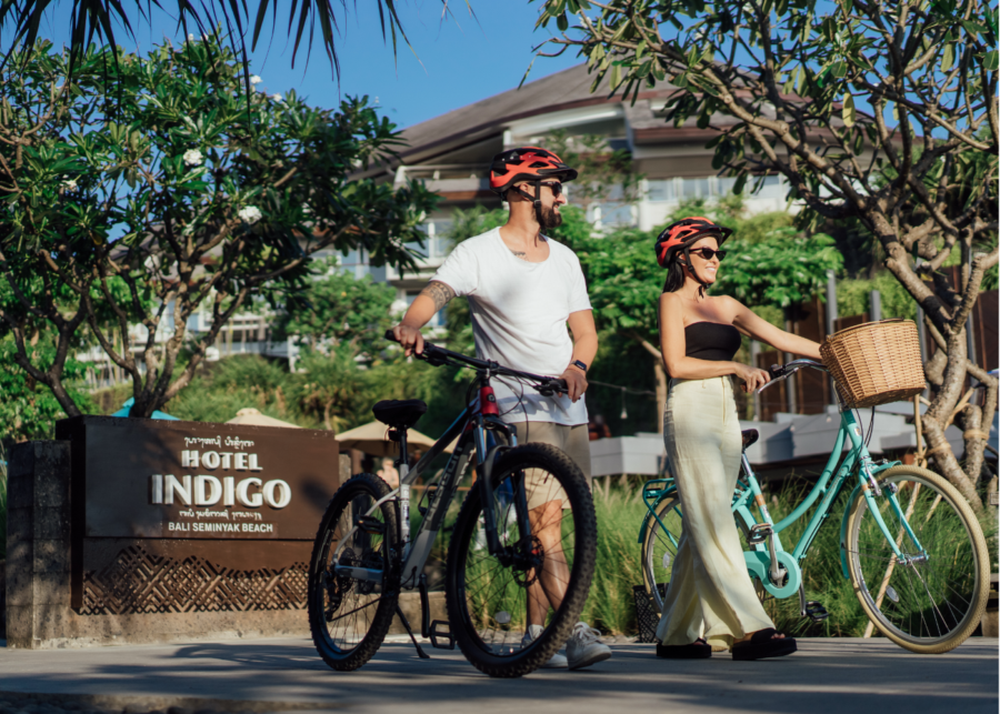 Two guests in red helmets walking bicycles in front of the Hotel Indigo Bali Seminyak Beach sign