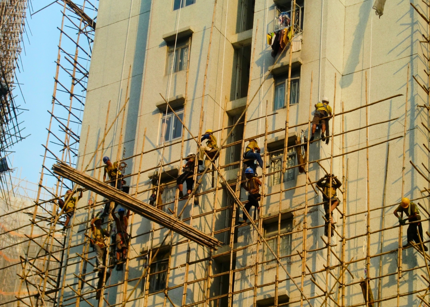Construction workers are working on bamboo scaffolding on the exterior of a building, with some wearing safety harnesses and helmets, suggesting a workplace safety investigation after an accident.